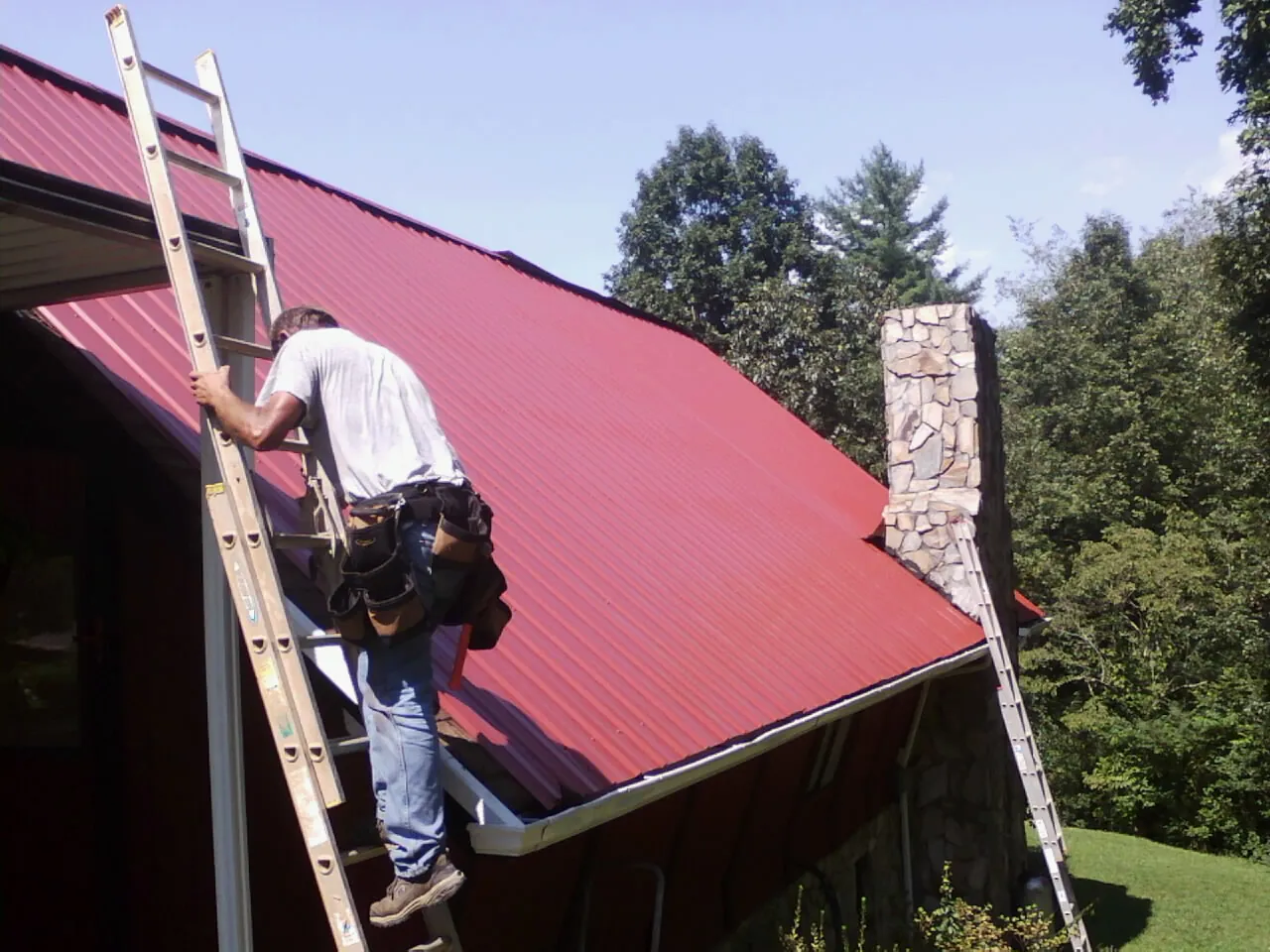 Roofer on a ladder beside a red metal roof and stone chimney — Soffit & Fascia Repair by Roofing Brothers in Clymer